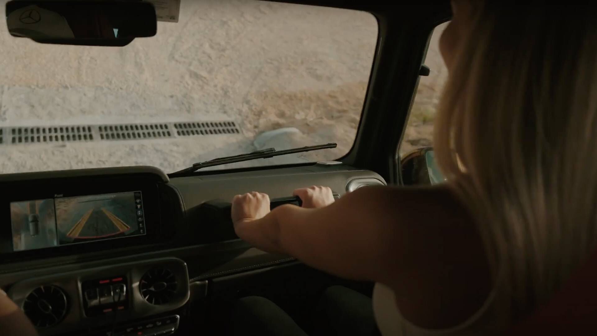 A blonde lady sitting inside a car holding onto a handle bar on a dashboard