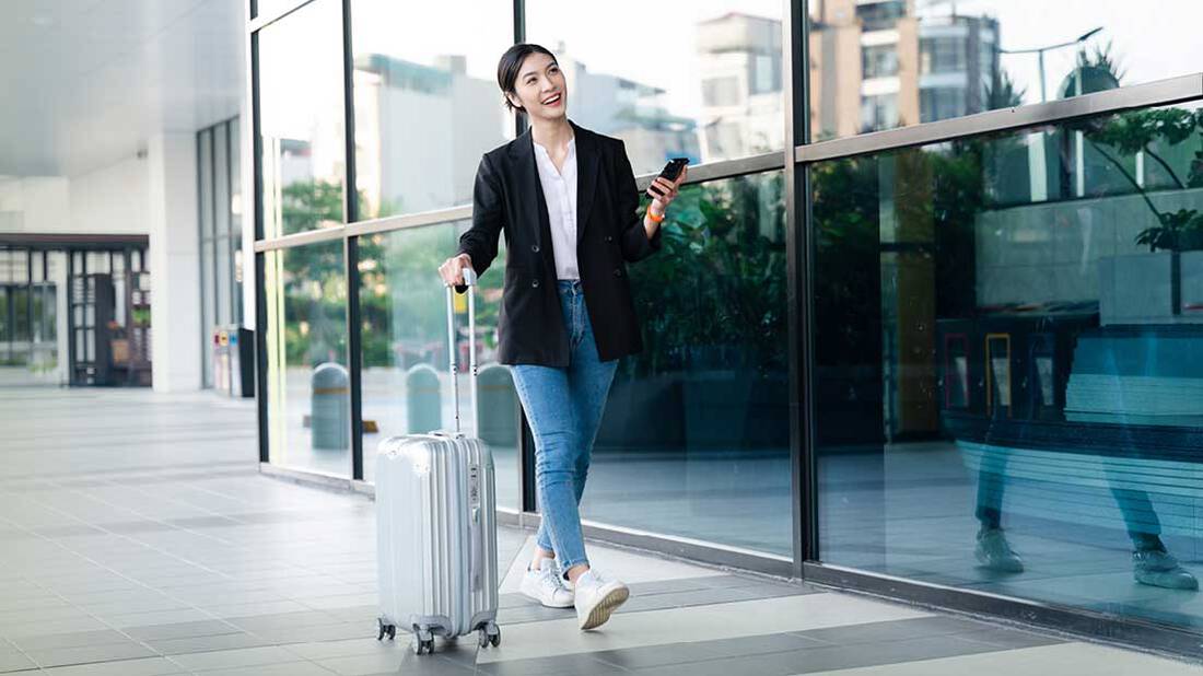 An Asian lady walking happily with a phone in one hand and luggage bag in the other