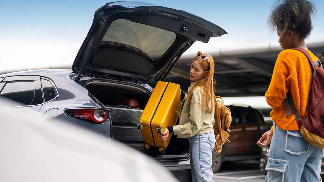 A blonde lady with backpack placing her luggage in the trunk of a car while another lady watches her